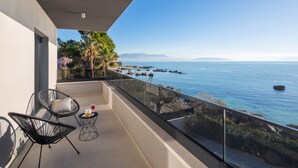 Un balcon avec deux chaises, une petite table et une balustrade en verre donnant sur une mer calme parsemée de rochers.