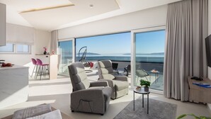 Un salon moderne avec un canapé, des fauteuils et une porte vitrée donnant sur un balcon d’où l’on peut admirer la mer.