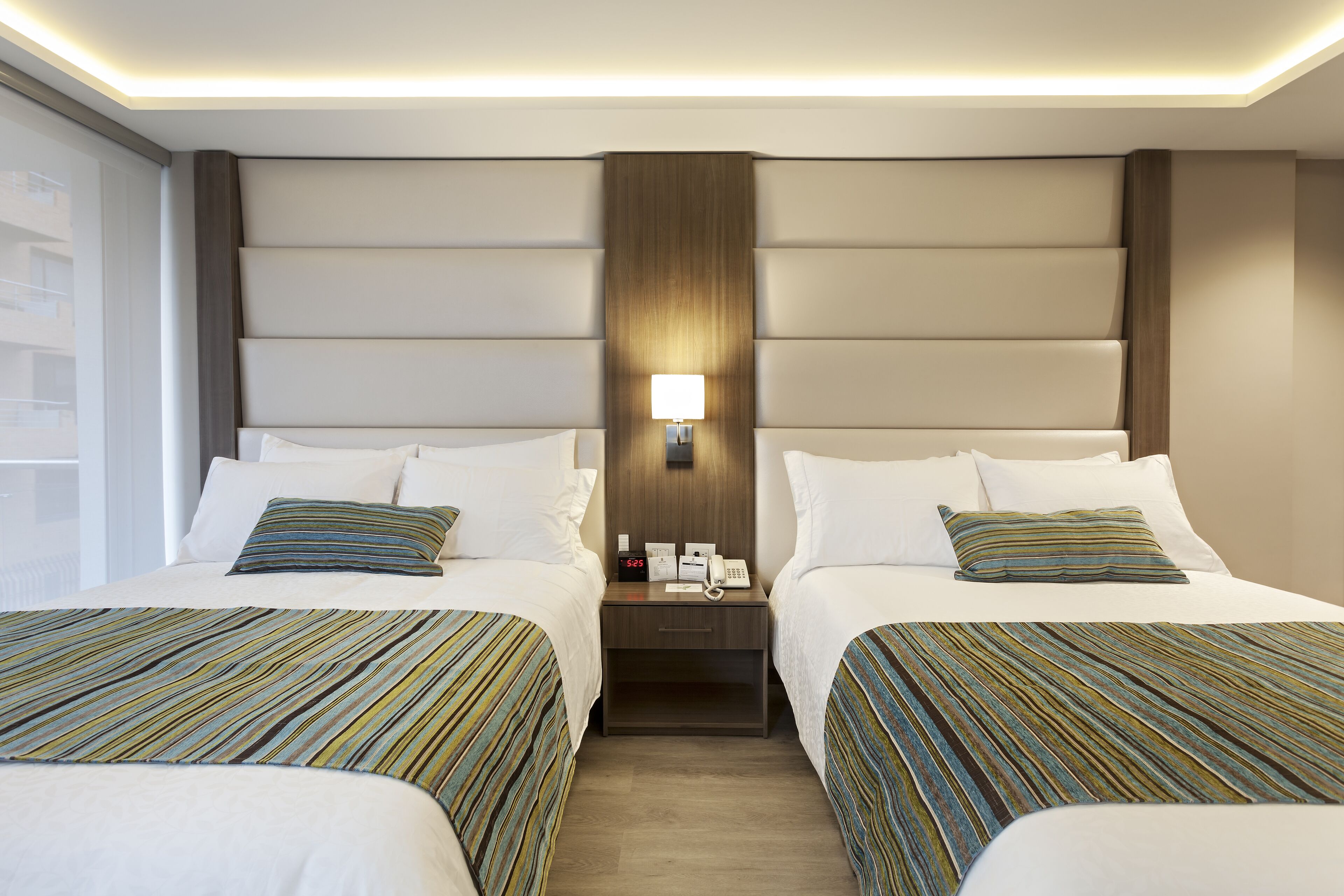 Habitación de hotel con dos camas, una mesita de noche con lámpara y vista al exterior.