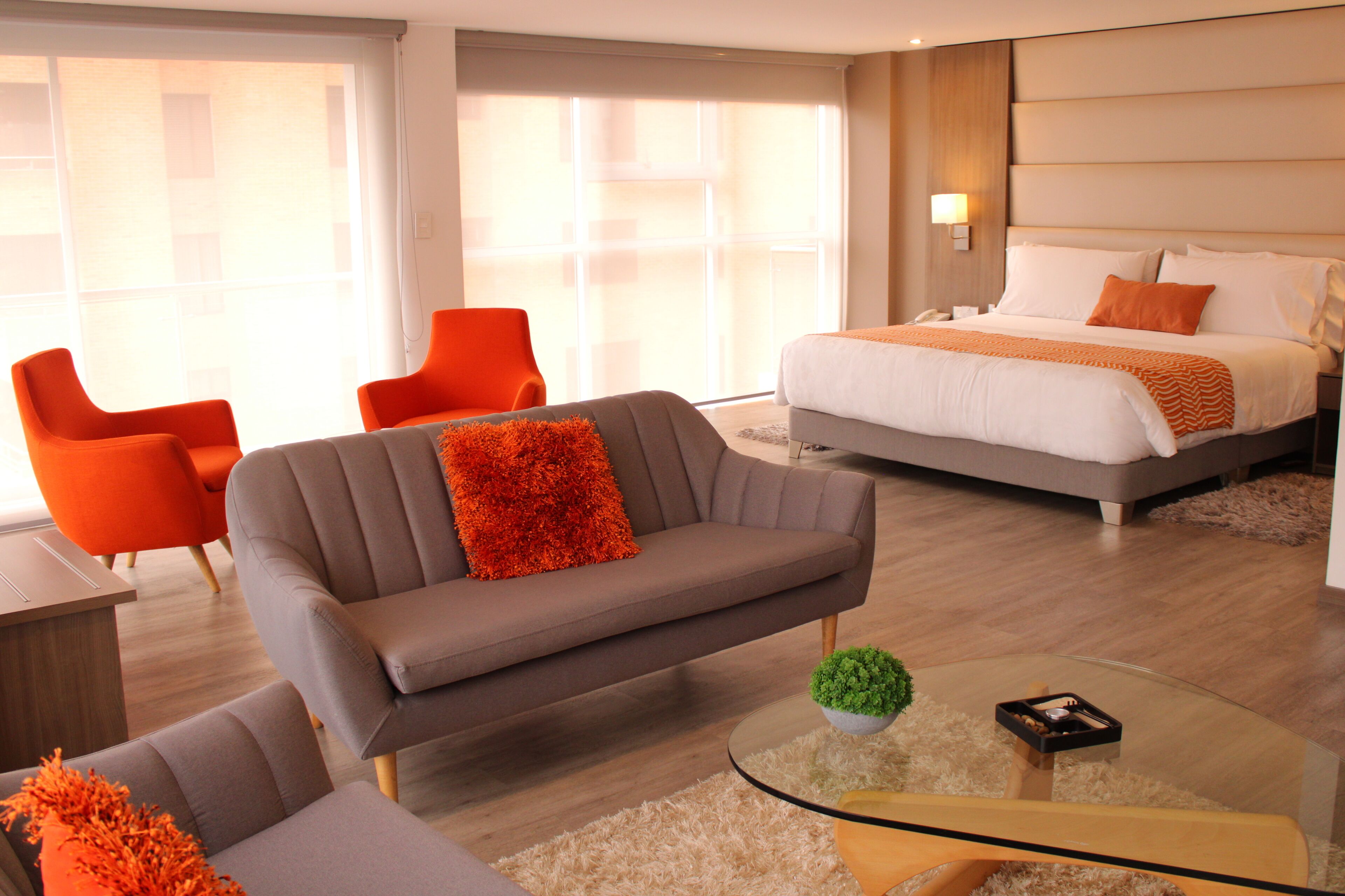Una habitación de hotel moderna con una cama, dos sillones naranjas, un sofá gris y una mesa de centro de vidrio.