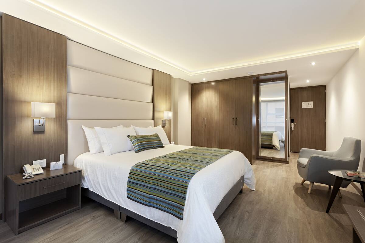 Ein modernes Hotelzimmer mit einem großen Bett, Nachttischlampen, einem Nachttisch, einem Schreibtisch mit Stuhl und einem Kleiderschrank.