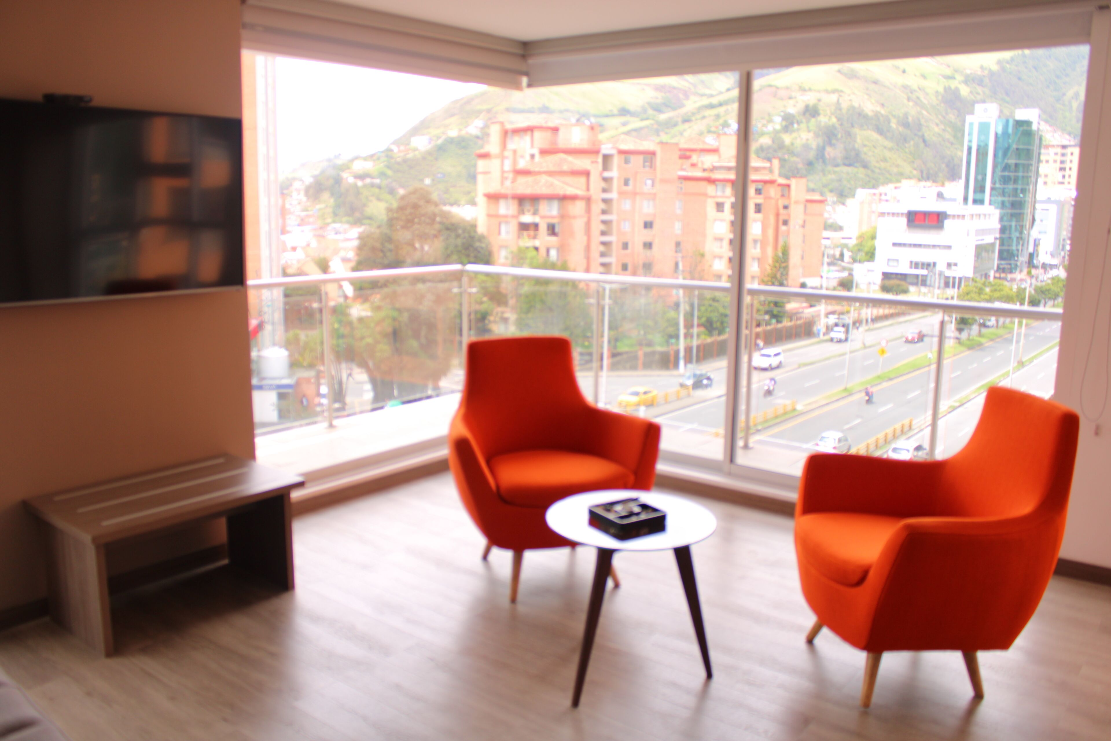 Una habitación moderna con dos sillas naranjas, una mesita y un ventanal amplio con vistas a la ciudad.