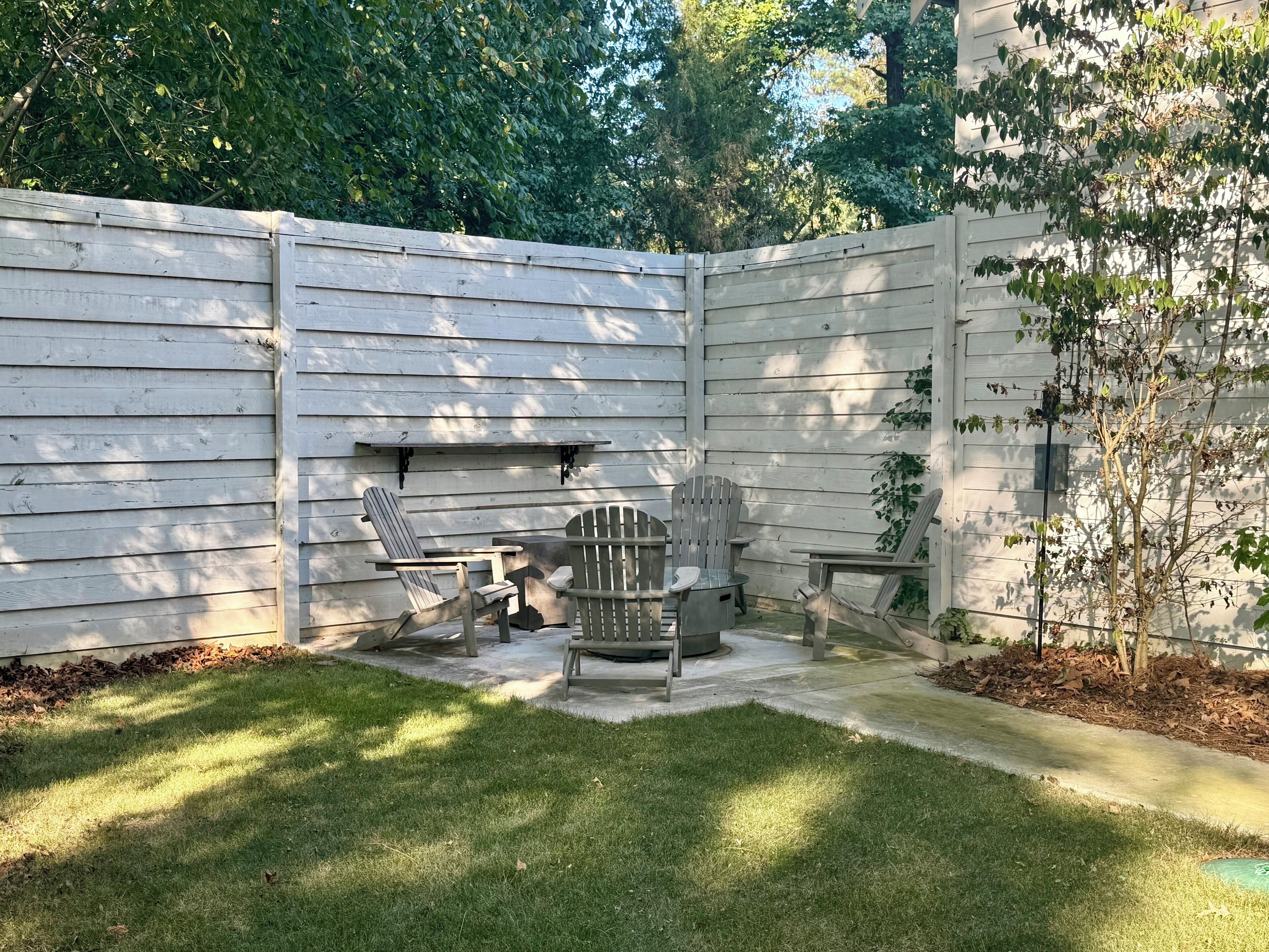 A backyard with a wooden fence, a concrete patio, and outdoor seating including two Adirondack chairs and a table.