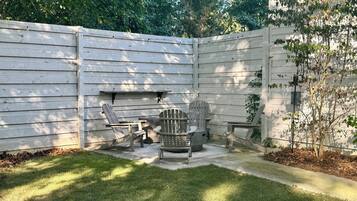 A backyard with a wooden fence, a concrete patio, and outdoor seating including two Adirondack chairs and a table.