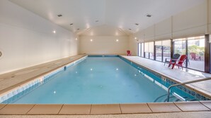 Una piscina interior con agua azul cristalina, bordes y suelo de baldosas. Hay luces en el techo y se ve una zona de estar exterior con sillas a través de amplias ventanas.