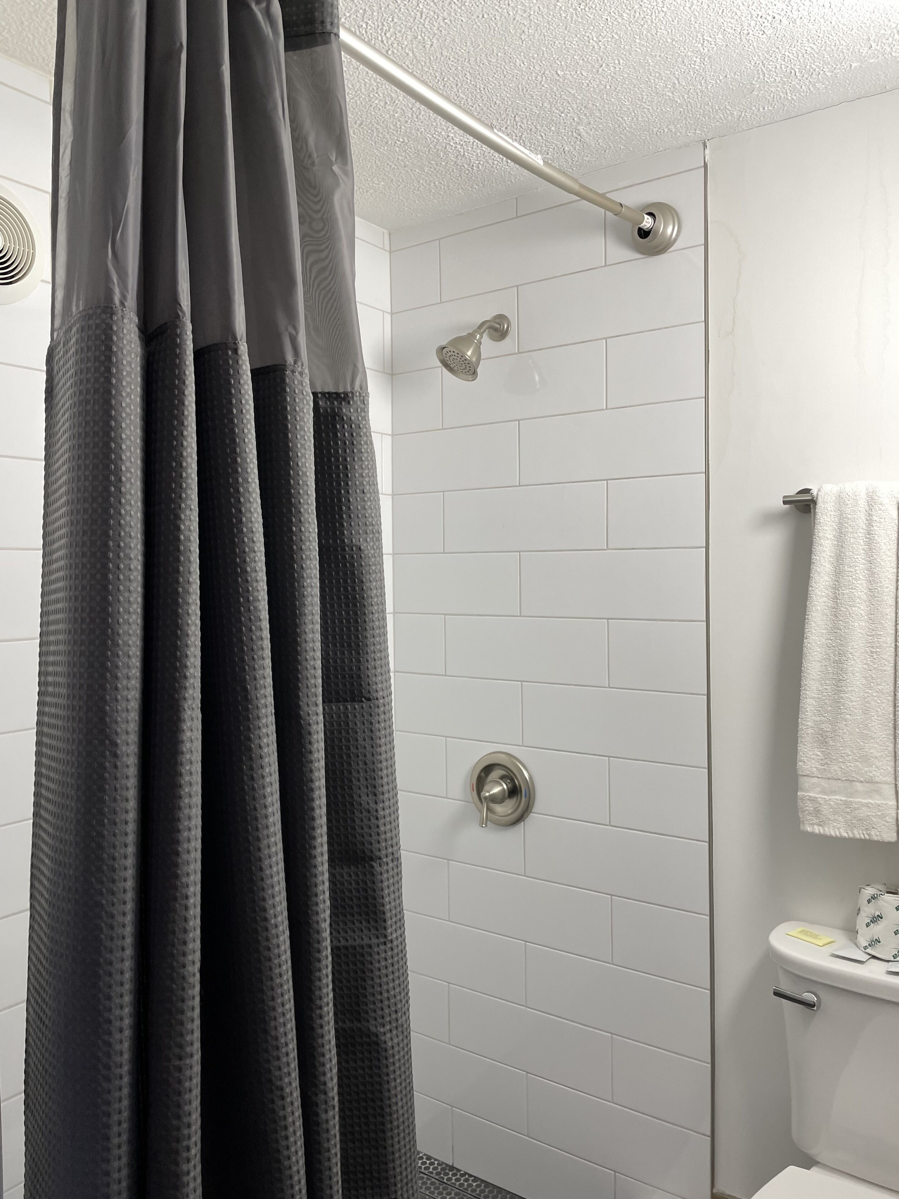 A shower area with a metal showerhead, a towel rack with a white towel, and a toilet with a paper towel dispenser.
