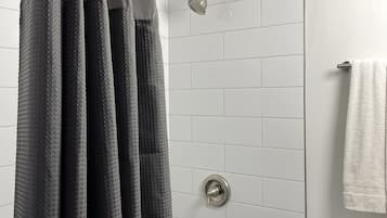 A shower area with a metal showerhead, a towel rack with a white towel, and a toilet with a paper towel dispenser.