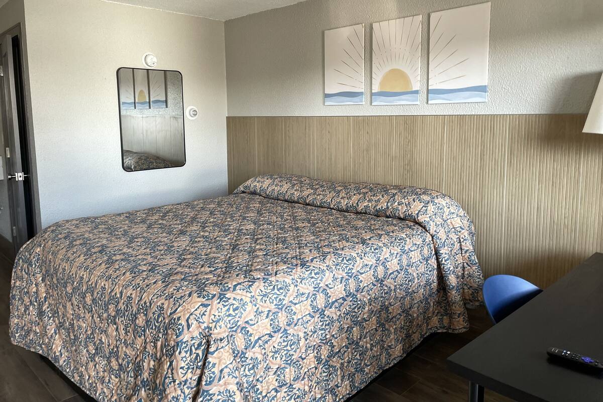 Un lit simple avec une couvre-lit à motifs, une table de chevet avec une télécommande, un miroir fixé au mur et un tableau représentant un coucher de soleil.