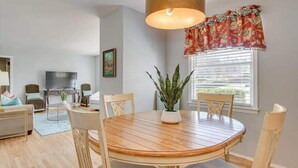 Een eethoek met een ronde houten tafel, vier stoelen, een potplant, een grote hanglamp en een raam met zonwering.