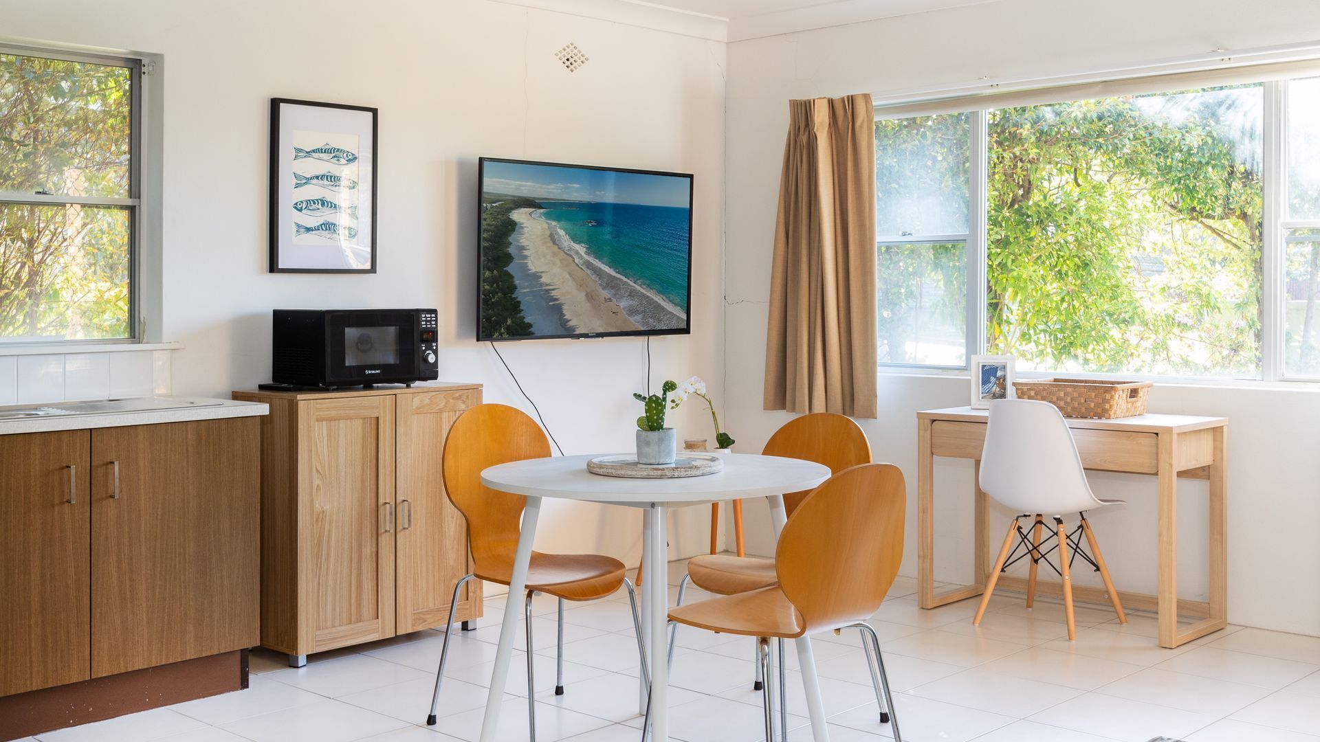 Ett modernt kök med mikrovågsugn, en matplats med ett runt bord och fyra stolar, en tv på väggen och utsikt över en strand genom fönstret.