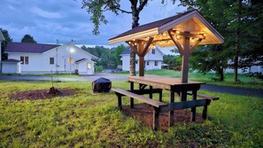 A wooden picnic shelter with benches, surrounded by grass and a dirt area, with houses in the background. - Hôtel à la maison - Chalet Lola (St-Tite)