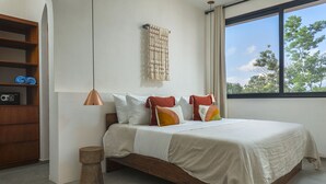 A bedroom with a bed, a wooden side table, a hanging light fixture, and a view of trees through a window. - Casa Ananta (Tulum)