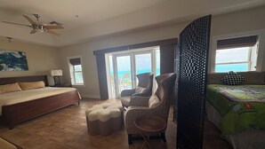 Een ruime slaapkamer met een groot bed, een zithoek met een fauteuil en voetenbankje, een plafondventilator en uitzicht op de oceaan door een schuifdeur.