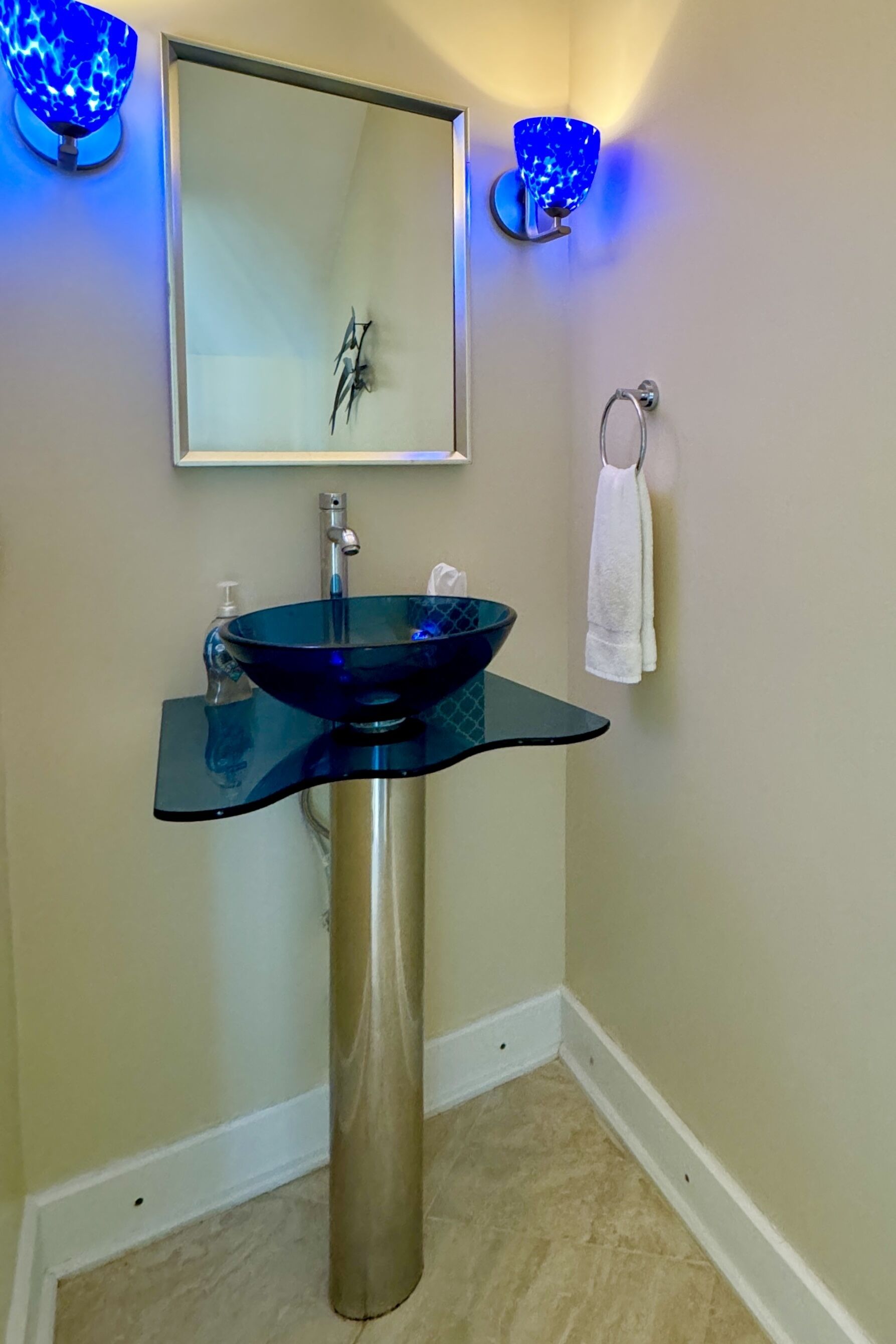 Un baño con un lavabo de vidrio, un espejo y dos apliques azules en la pared.