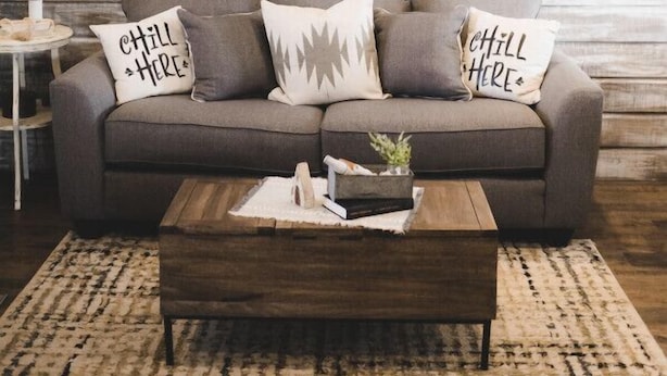 A living room with a grey sofa, multiple cushions, a wooden coffee table, and a rustic wooden wall.