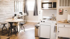 A compact kitchen with a stove, microwave, and sink. There is a dining table with chairs and a small countertop with a kettle and plates.