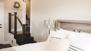 A bedroom with a bed, white bedding, a wooden headboard, a nightstand, a lamp, and a staircase.