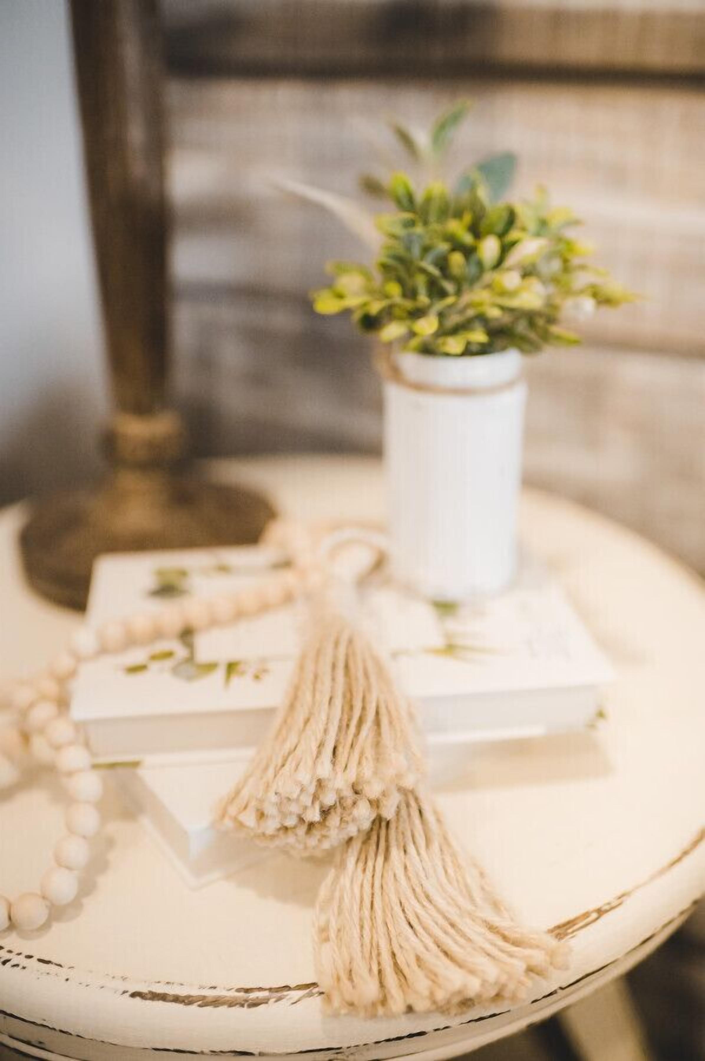 Une table ronde blanche, à l’aspect vieilli, agrémentée d’une petite plante en pot, d’un collier de perles et d’une frange.