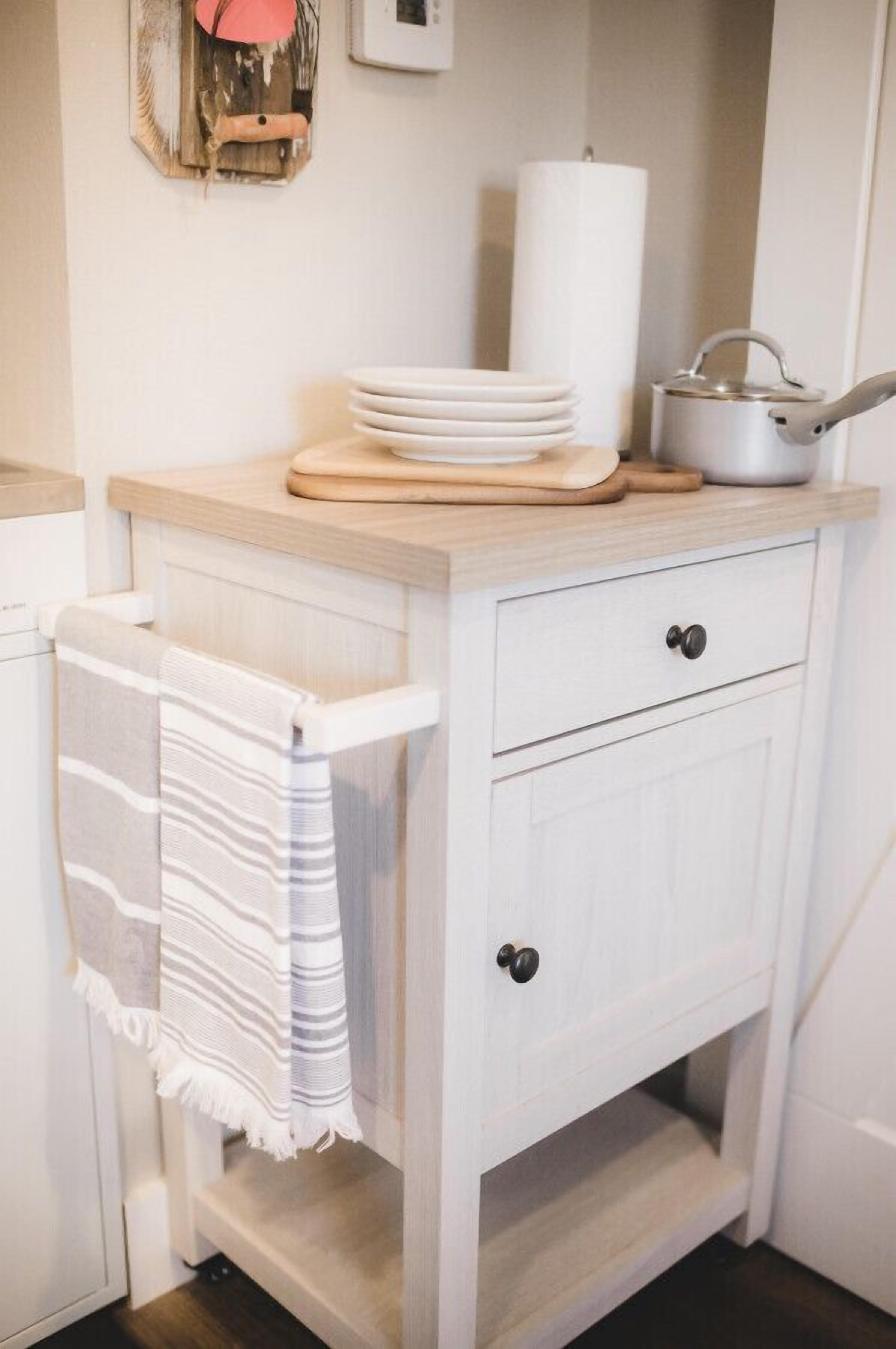 Une cuisine avec un meuble blanc, une serviette accrochée à la porte, une pile d’assiettes blanches, une planche à découper, une bougie blanche et une casserole posée sur le comptoir.