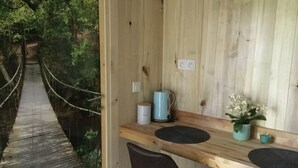 Una cabaña de madera con una mesa y sillas de madera, una tetera y una pequeña planta. Un puente colgante conduce a la cabaña.