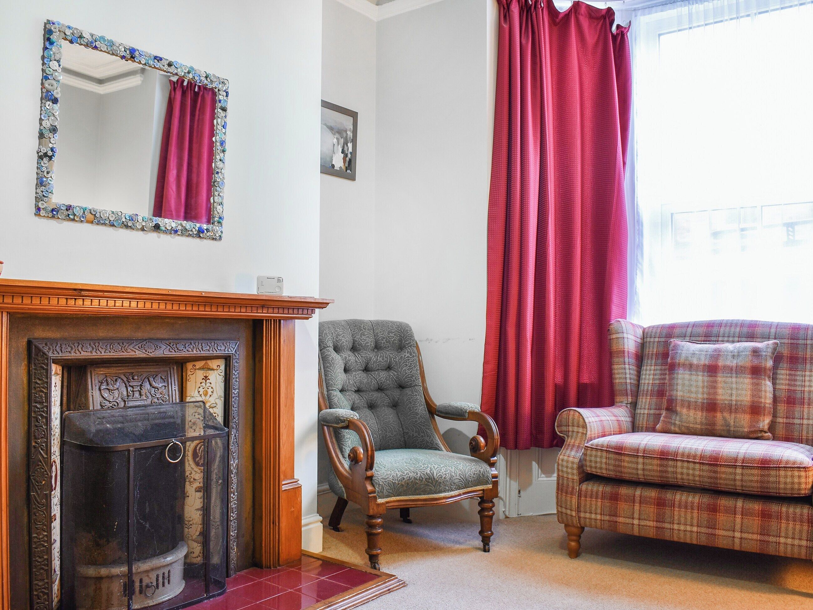A room with a fireplace, a mirror, a green armchair, a plaid sofa, and a large window with red curtains.