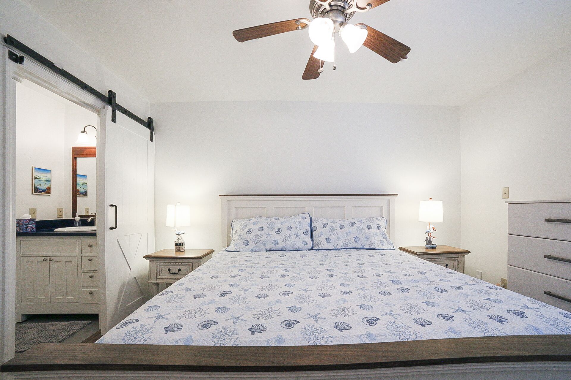 Une chambre à coucher avec un lit, deux tables de chevet avec des lampes, un ventilateur de plafond, une commode et une salle de bain visible à travers une porte ouverte.