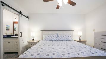 A bedroom with a bed, two bedside tables with lamps, a ceiling fan, a dresser, and a bathroom visible through an open door.
