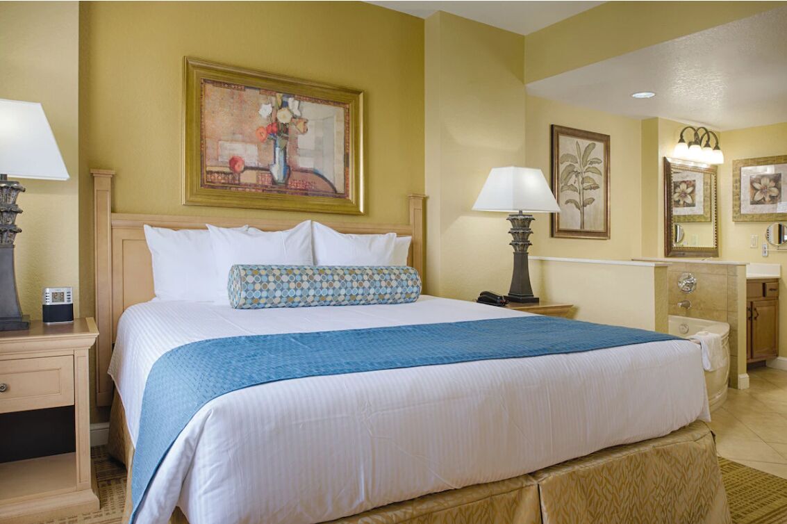 A hotel room with a large bed, a nightstand with a lamp, a bedside table, a bathroom with a sink and mirror, and a painting on the wall.