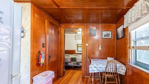 A compact cabin with wooden walls, a small dining area with a table and chairs, a visible bed, and a fire extinguisher. - Gorgeous Escape, Steps Away from Fourth Lake at Stiefvater's Lakeside Cottages (Inlet)