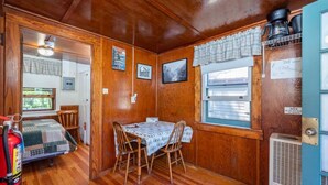A compact room with wooden walls and a small dining table with chairs. A fire extinguisher is visible on the left, and a window with a curtain is on the right. - Gorgeous Escape, Steps Away from Fourth Lake at Stiefvater's Lakeside Cottages (Inlet)