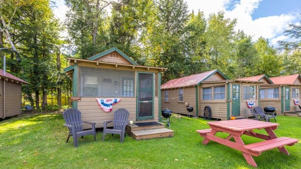 A row of cabins with a picnic table and chairs in the foreground, surrounded by a grassy area and trees. - Gorgeous Escape, Steps Away from Fourth Lake at Stiefvater's Lakeside Cottages (Inlet)