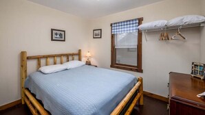 Un dormitorio con una cama de madera, una mesita de noche con lámpara, una ventana con persianas y una estantería con percheros.