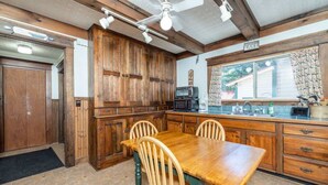 A kitchen with wooden cabinets, a dining table with chairs, a microwave, and a window with a view of a house. - Lakeside House View Overlooking Beautiful 4th Lake Here at Stiefvater's Lakeside Cottages (Inlet)