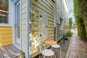 Une terrasse en bois avec deux chaises en métal blanc et une petite table, une porte blanche et, au fond, une clôture en bambou.