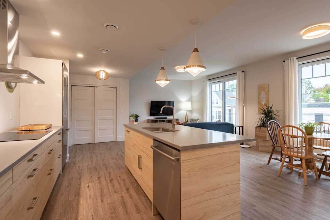 Een moderne keuken met een centraal eiland, houten kastjes en een vaatwasser. Er is een zithoek met een tafel en stoelen, een televisie en een raam met gordijnen.