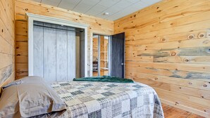 Une chambre avec des murs en bois, un lit recouvert d’une courtepointe, un plaid vert et une porte donnant sur une autre pièce.