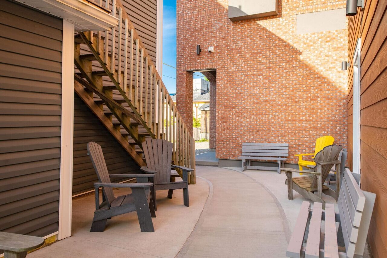 A narrow outdoor corridor with wooden steps, a brick wall, and outdoor seating including Adirondack chairs and a bench.