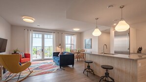 A modern living room with a sofa, a rocking chair, a dining area with a table and chairs, and a kitchen with a bar counter and stools. - Suite 304 - 470 Main (Wolfville)