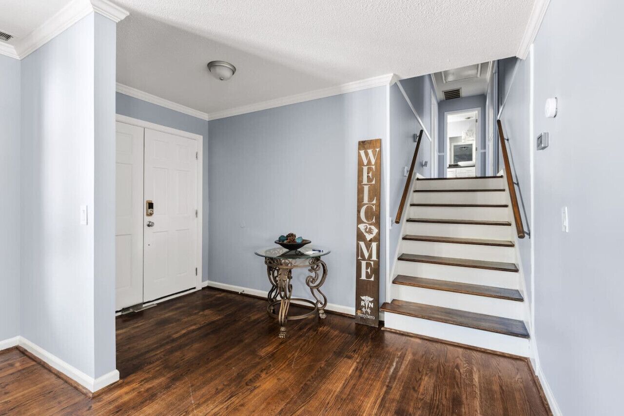 A hallway with a wooden floor, a white door, a glass table with a decorative bowl, and a wooden staircase with a 'WELCOME' sign.