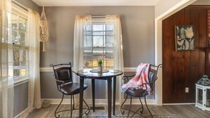 Una habitación con una mesa, dos sillas, una ventana con cortinas, un radiador y una puerta de madera con una planta y un farol.