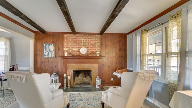 Una habitación con chimenea, dos sillones, un tapiz estampado, vigas de madera y una ventana con cortinas.
