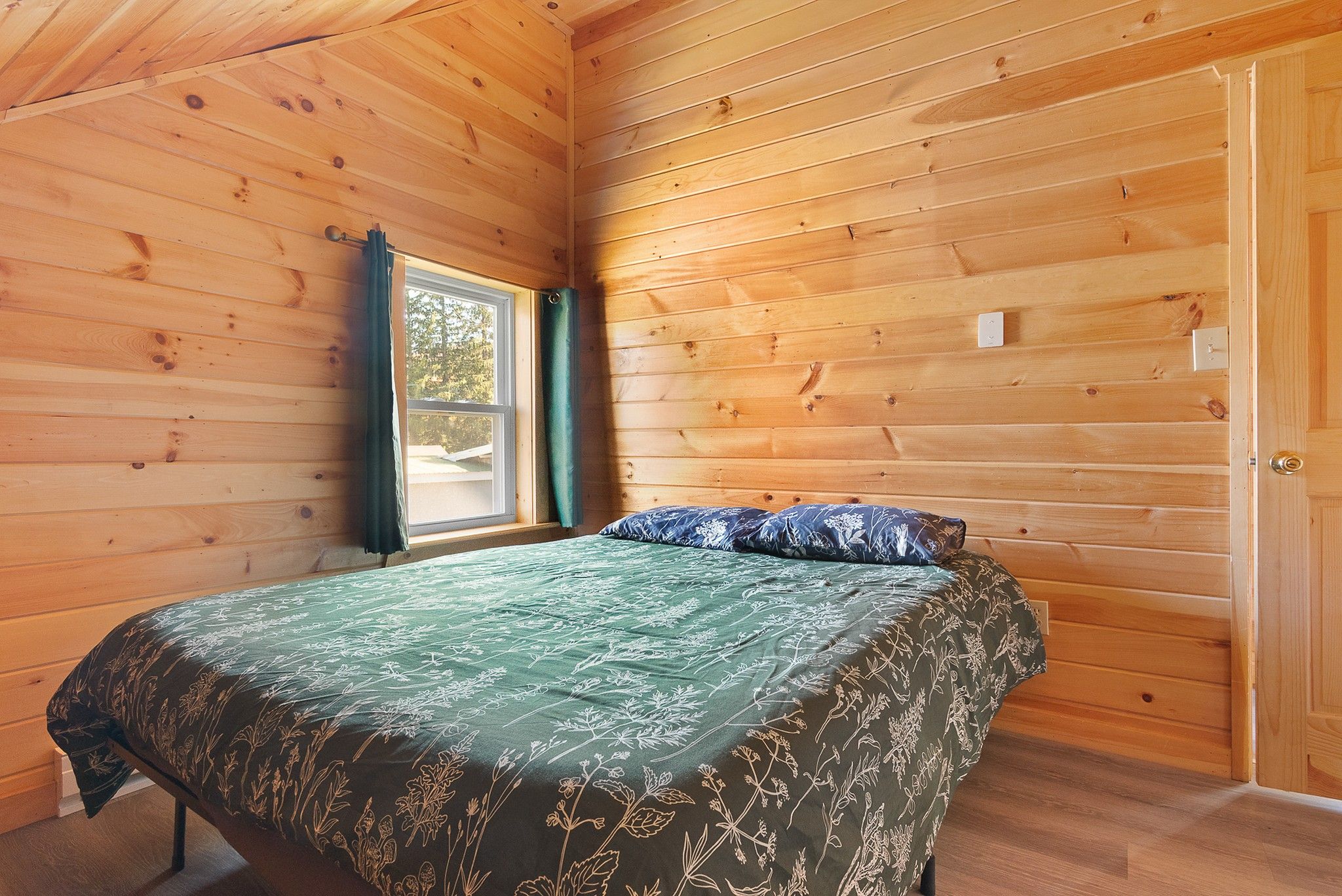 A wooden room with a bed, a window with curtains, and a door.