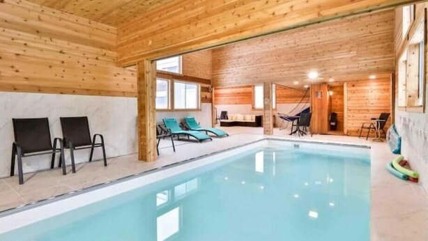 An indoor swimming pool with wooden walls and ceiling, surrounded by lounge chairs and a hammock.