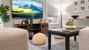 Uma sala de estar com televisão de tela plana, uma luminária, uma estante de livros e um tapete estampado.