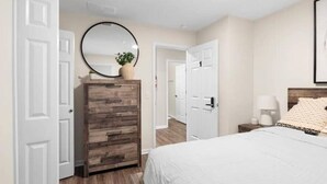 Um quarto com cômoda de madeira, espelho redondo, cama com roupa de cama branca e mesa de cabeceira com abajur.