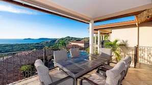 A terrace with a table and chairs, a pool, and a view of the sea and hills. - Bay Point 7 • 3 BR Ocean-View Condo • Two Infinity Pools, Playa Panamá - 6 Guests (Playa Hermosa)