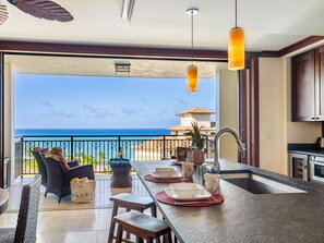 A kitchen with a view of the ocean, a dining area with chairs and a table, and a bar area with stools. - Hale Keani O-1203 stunning Views to relax: Special book 7 NIGHTS in September AND GET 1 NIGHT FREE (Kapolei)