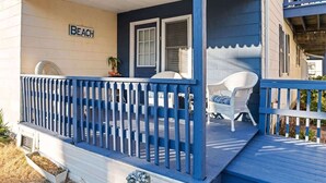 Un porche couvert avec une balustrade bleue, une enseigne indiquant « PLAGE » et deux chaises en osier blanches.