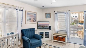 Un salon avec un fauteuil bleu, un téléviseur posé sur un meuble, un ventilateur de plafond et une vue par la fenêtre.