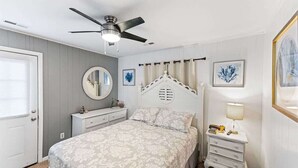 Un dormitorio con una cama, una mesita de noche, un ventilador de techo, un espejo y una puerta.